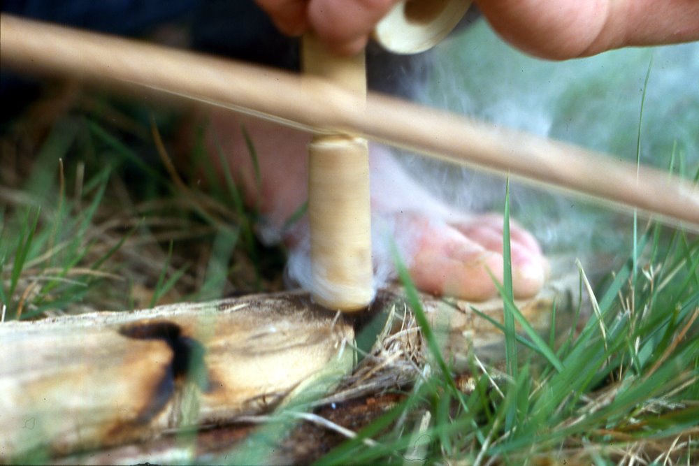 Bow drill kindling a need fire by friction, smoke rising from the hearth board